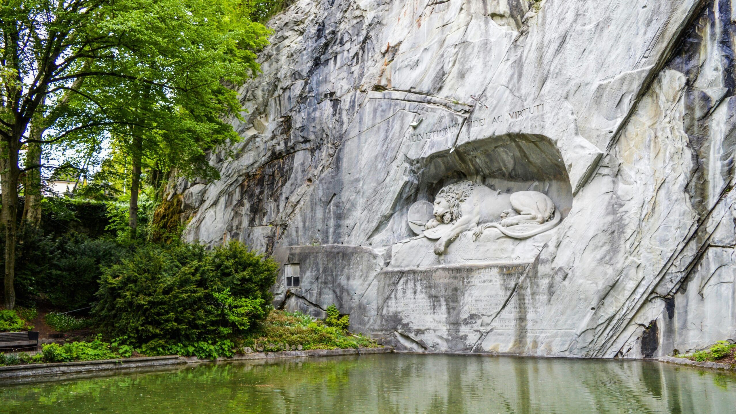 Sculpture of a mortally wounded lion carved into sheer rock ledge near pond located in Switzerland