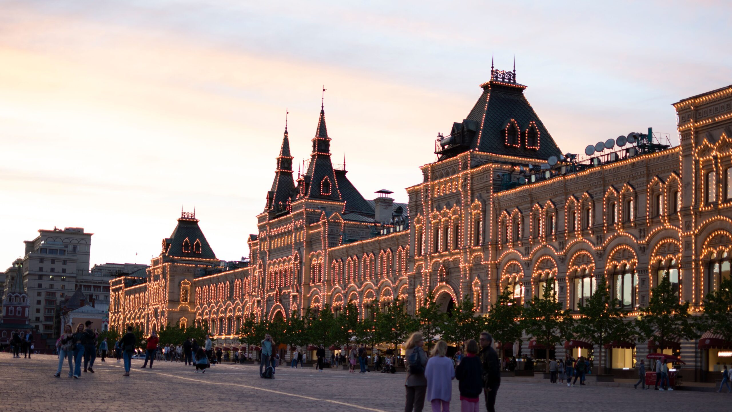 View of GUM Department Store lit up at twilight in Moscow's Red Square, with people strolling in the foreground.