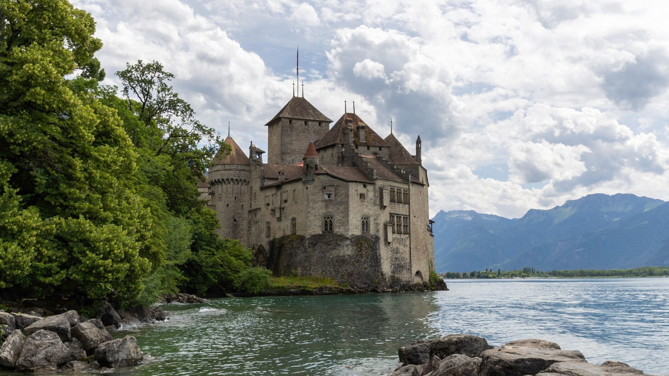 Scenic view of Château de Chillon by the shores of Lake Geneva, surrounded by mountains and lush greenery.